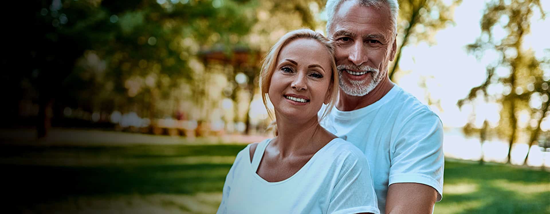 Smiling middle-aged couple in light blue shirts outdoors in a park, representing ongoing aesthetic and wellness care.