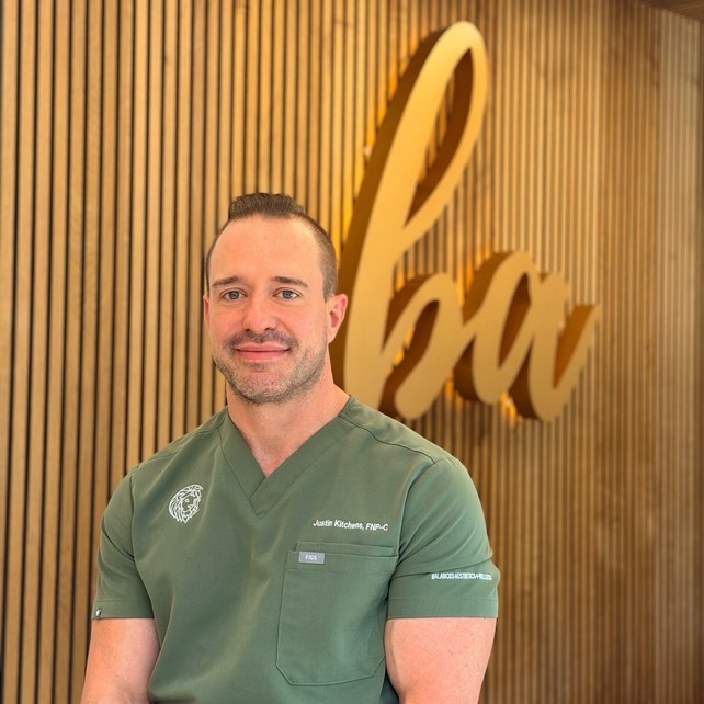 A man withJustin Kitchens, FNP-C — Lead Wellness Clinician at Balanced Aesthetics + Wellness short hair and a trimmed beard is standing in front of white curtains, wearing a black short-sleeve scrub top and smiling slightly at the camera.